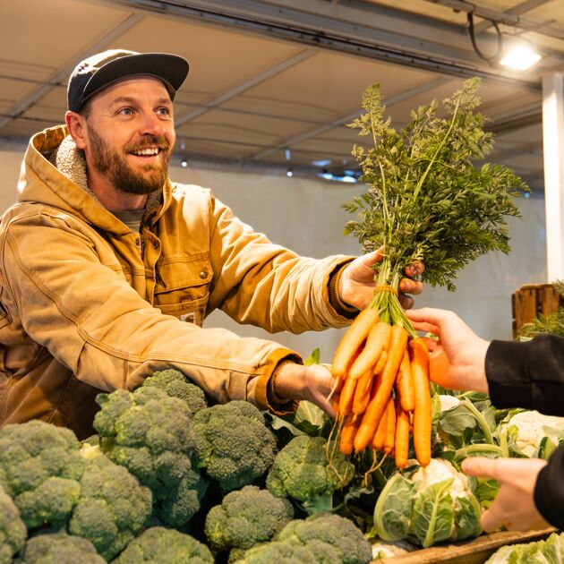 Lachende man met pet geeft wortels aan iemand in een kraam. 
