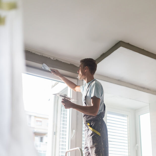 Man in een overall op de ladder is het plafond aan het stuken.