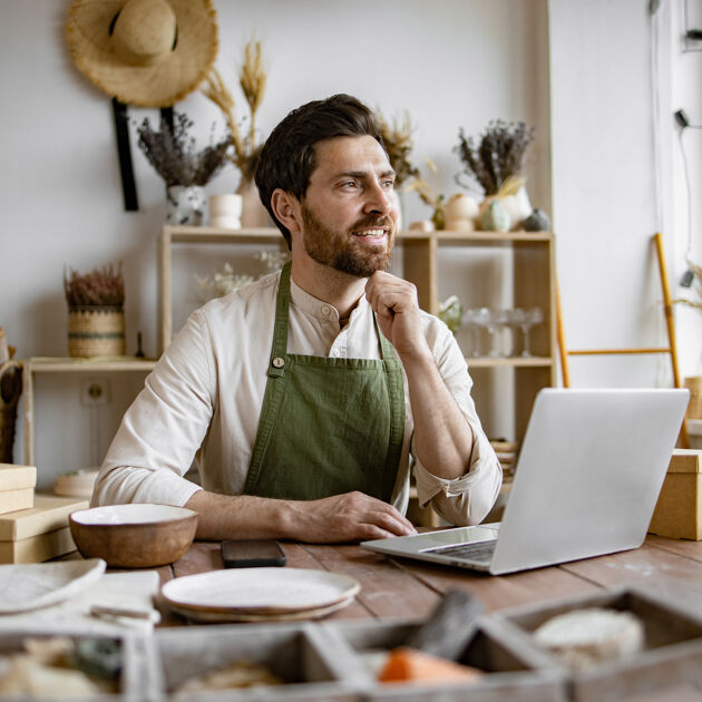 Man met een schort zit aan een rommelig bureau en kijkt dromend weg van zijn laptop. 