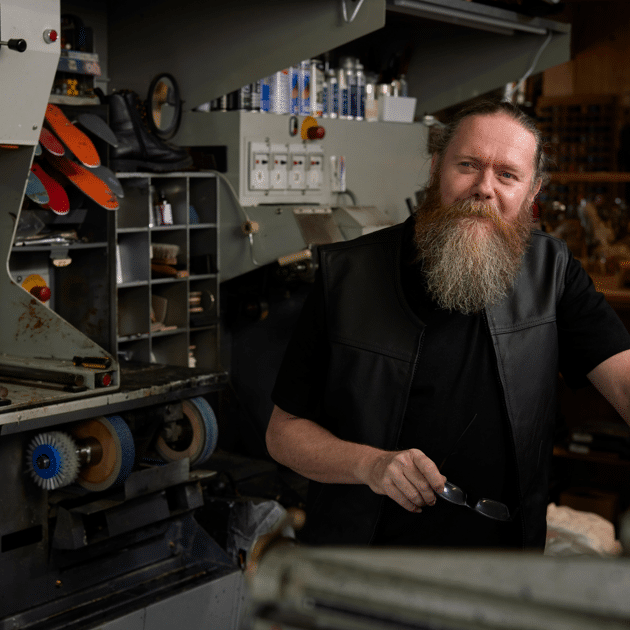 Man met baard in zwarte kleding zit naast machines voor het maken van schoenen
