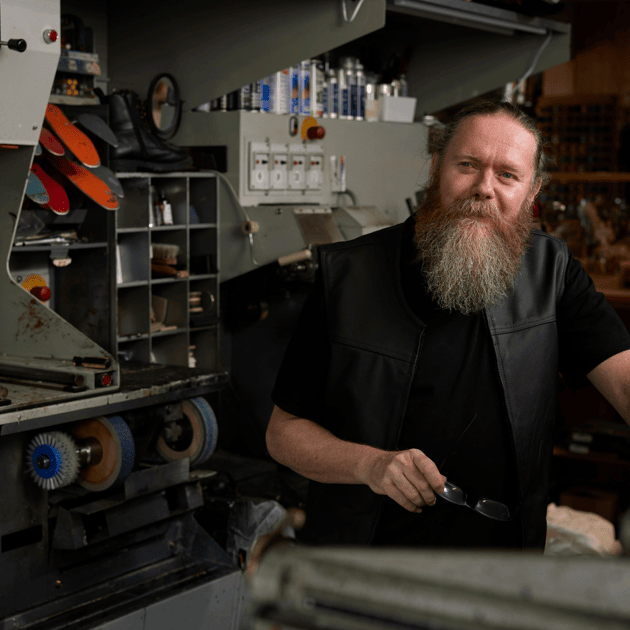 Man met baard in zwarte kleding zit naast machines voor het maken van schoenen