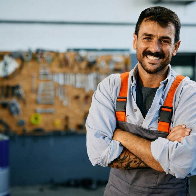 Lachende man met overall staat lachend met zijn handen over elkaar voor een muur met gereedschap.