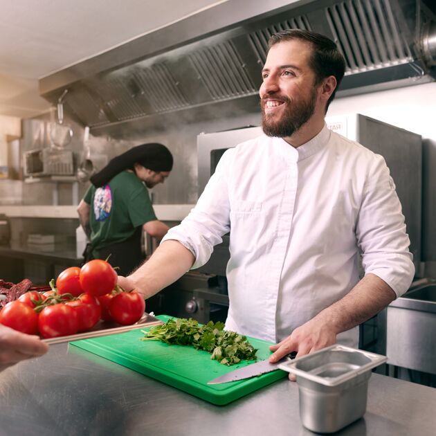 Man staat in de keuken en lacht terwijl hij tomaten van iemand aanneemt. 