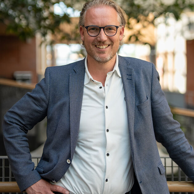 Man in pak met bril leunt buiten tegen hek aan