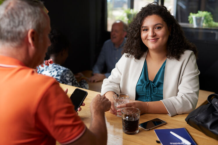 Vrouw in wit jasje lacht naar camera terwijl ze aan tafel zit bij training