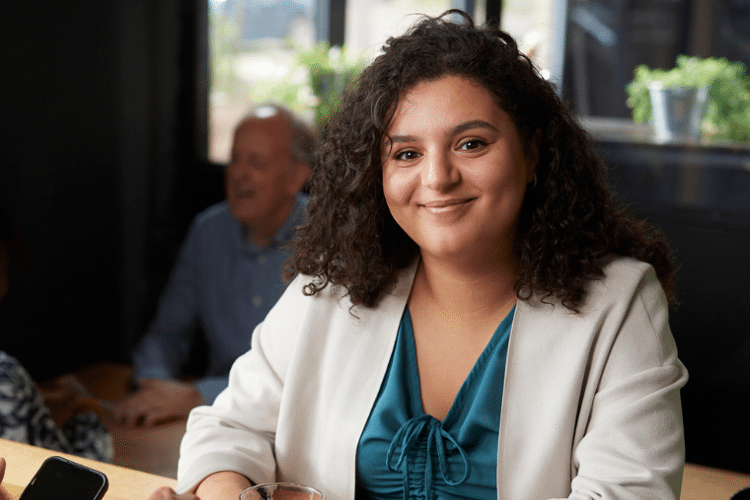 Vrouw in wit jasje lacht naar camera terwijl ze aan tafel zit bij training