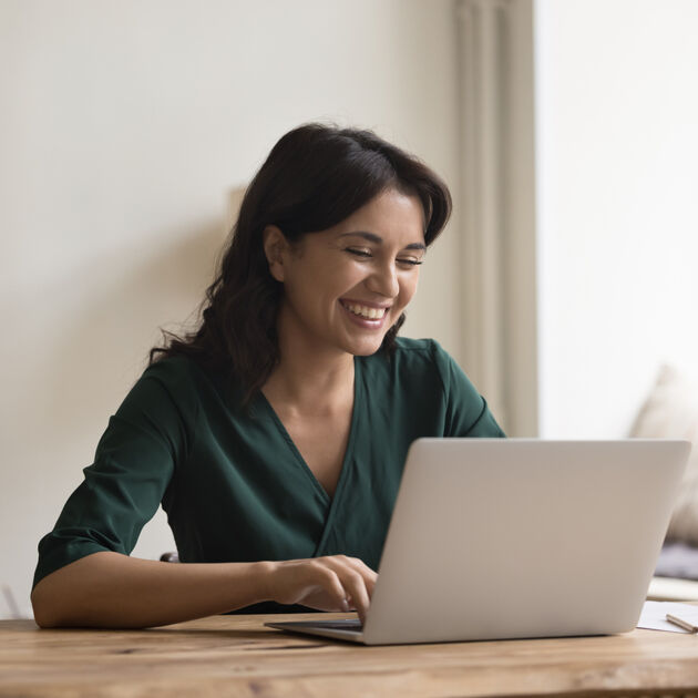 Vrouw zit aan tafel en kijkt lachend naar laptop.