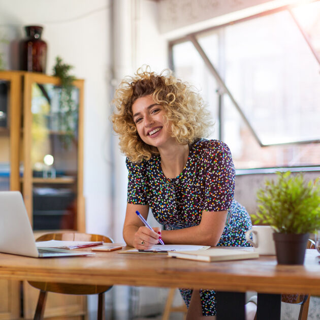 Lachende vrouw met krullend haar staat achter bureau met pen in haar hand. 