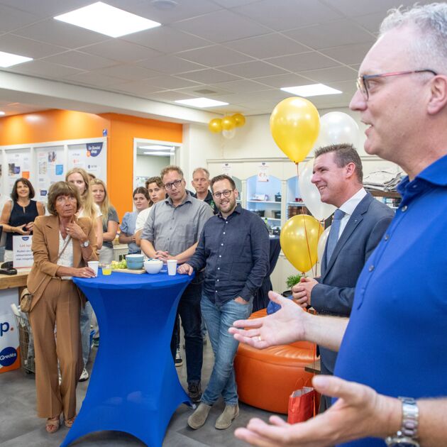 Een groep mensen bij een blauwe tafel kijken naar een man in een blauw shirt. Er zijn gele ballonnen.