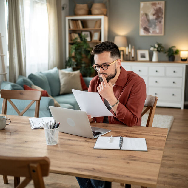 Een man zit aan tafel met laptop en kijkt bedenkelijk naar papier.