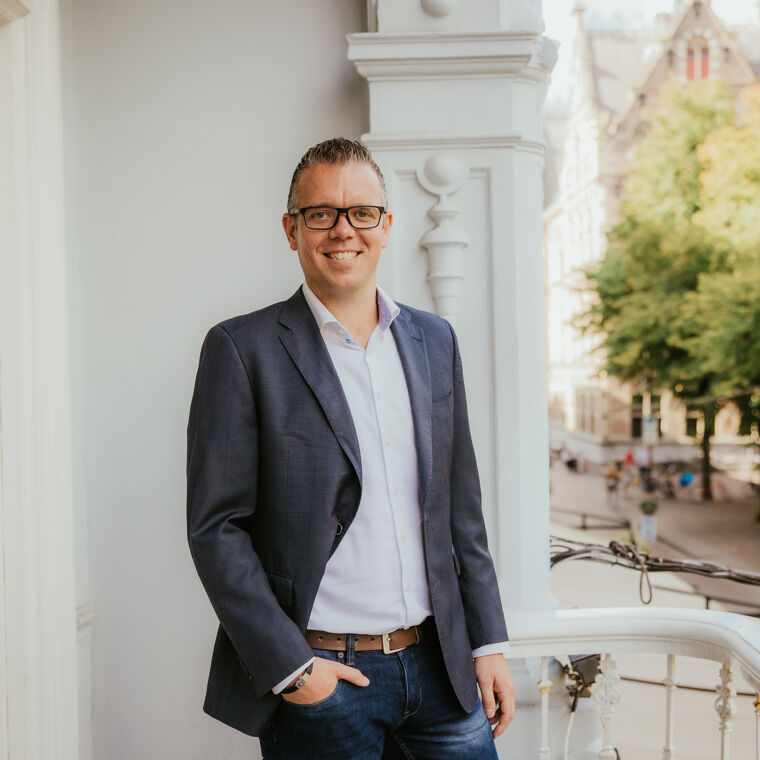 Man in pak met bril kijkt naar de camera terwijl hij lacht.