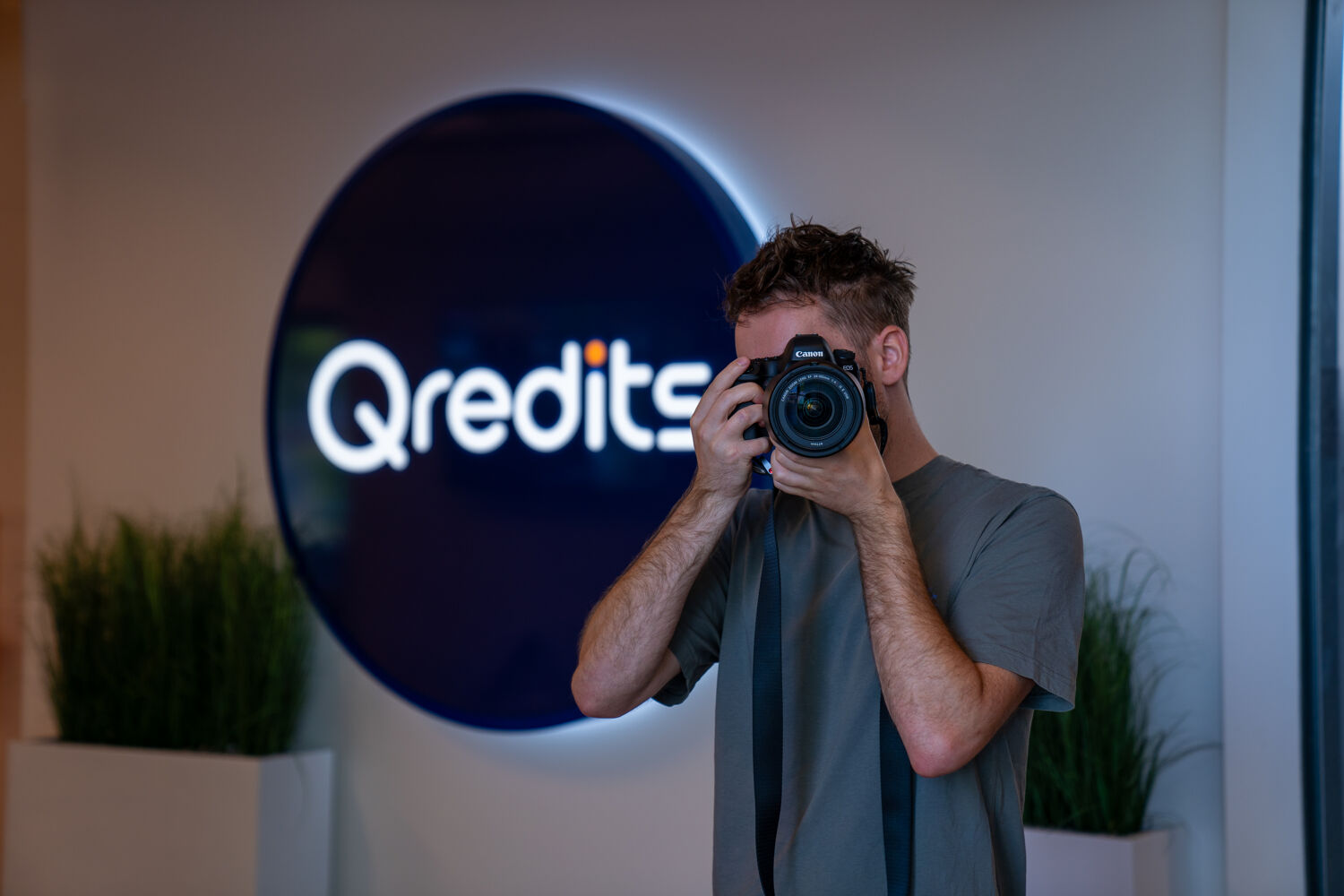Man in groen shirt staat voor een blauw Qredits logo met een camera in zijn handen