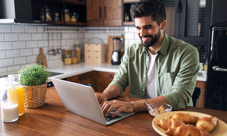 Een man zit in de keuken, lachend achter zijn laptop.