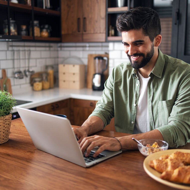 Een man zit in de keuken, lachend achter zijn laptop.