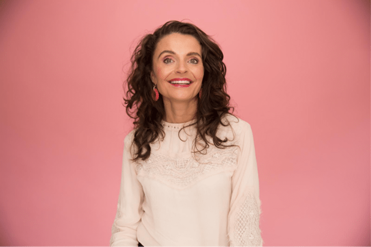 Vrouw met wit shirt en bruine haren, lachend voor roze muur.

