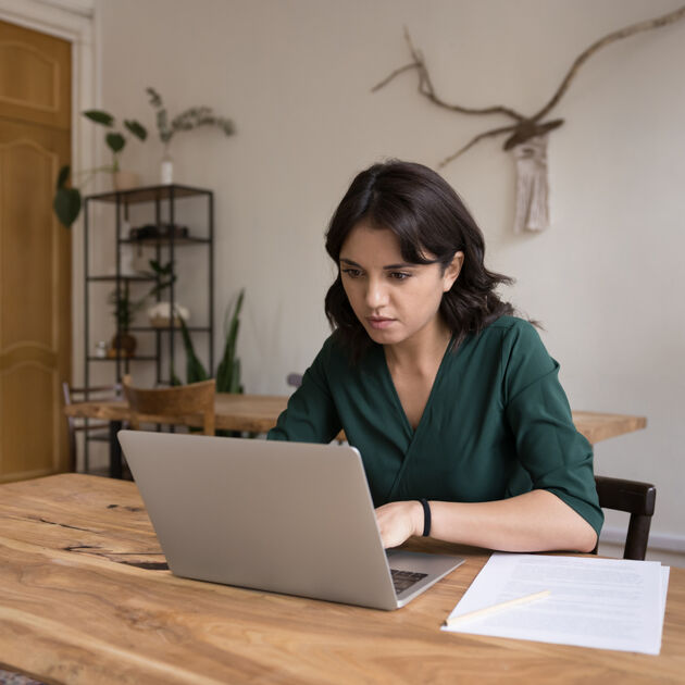 Vrouw kijkt serieus en zit aan tafel werkend achter laptop.