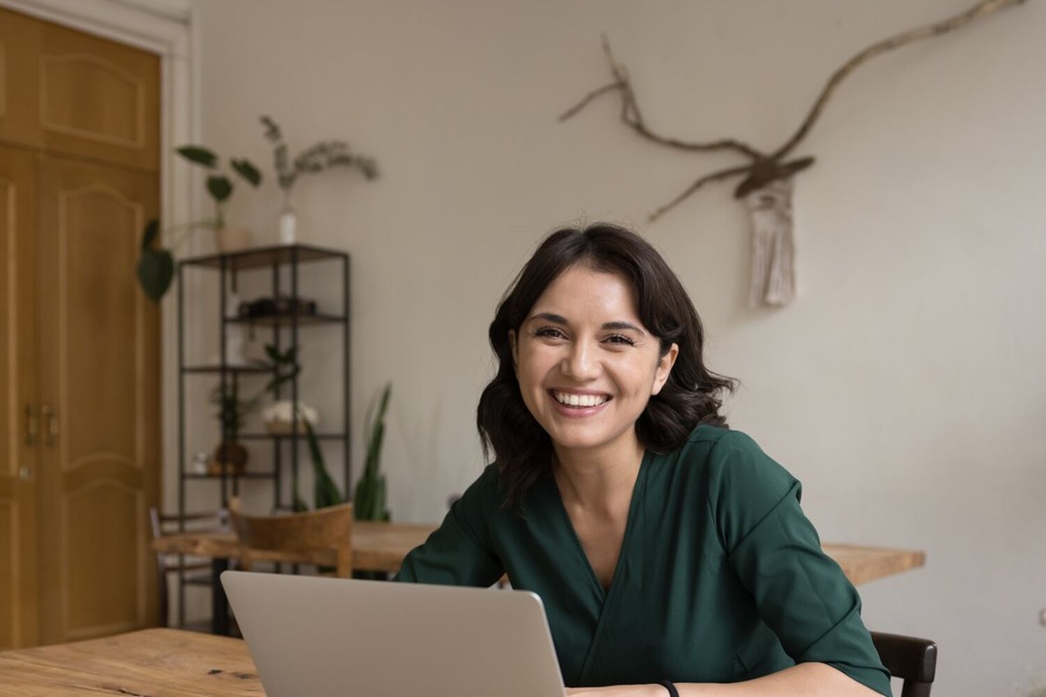 Vrouw zit aan tafel achter laptop, kijkt lachend.