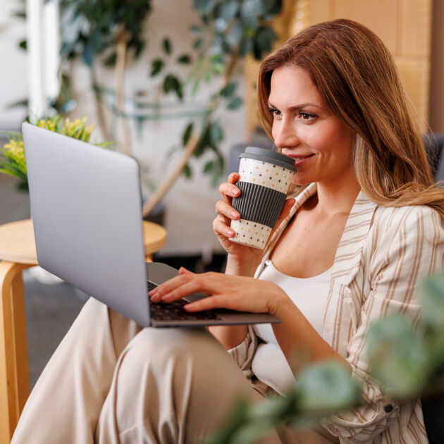 Vrouw met een koffiebeker in haar hand en laptop op schoot kijkt lachend.